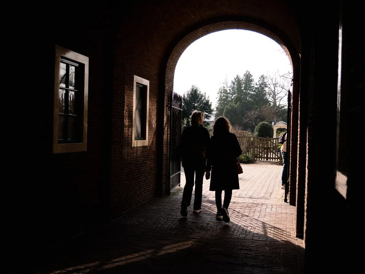 Two figures silhouetted in a dark brick archway that opens onto a sunlit garden path