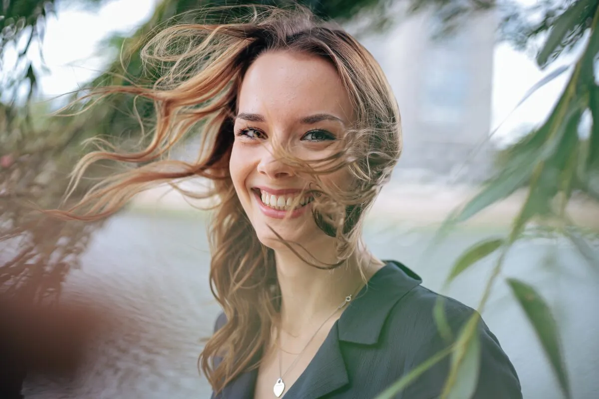 Woman laughing outdoors with her hair blowing across her face, framed by soft green leaves and water behind her