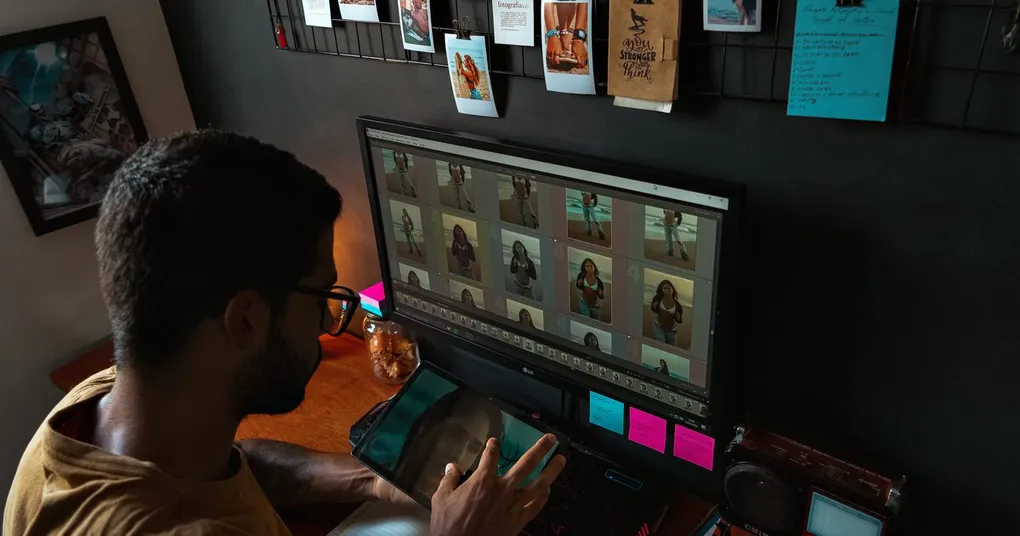 Photographer at a desk reviewing a grid of portrait thumbnails in photo editing software on a monitor, with prints pinned on the wall above