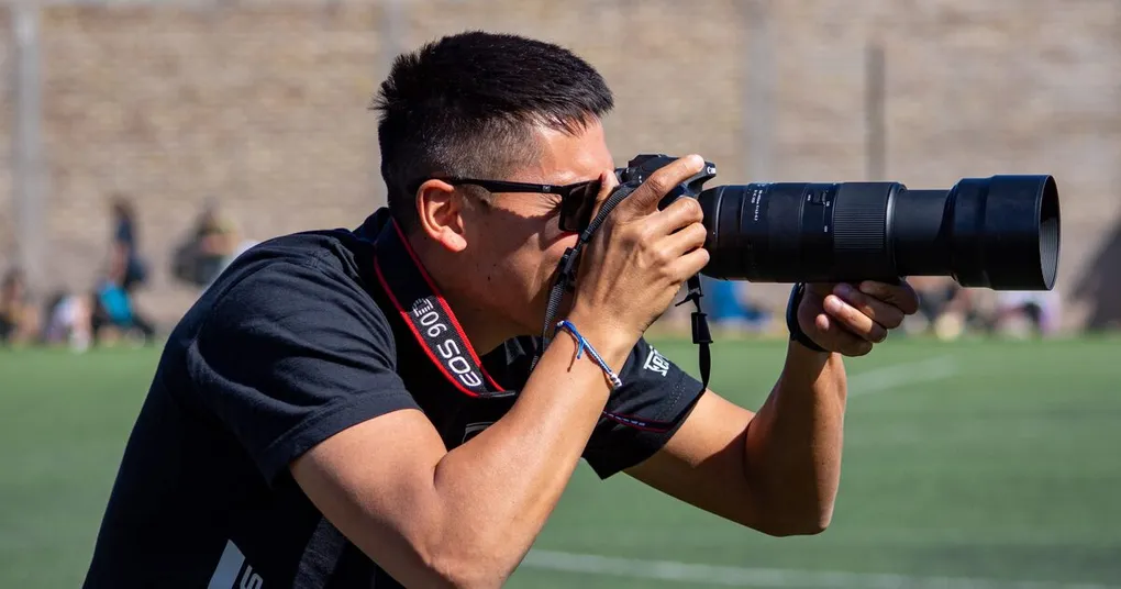 Sports photography in progress: a photographer in profile shooting across a green pitch with a DSLR and long telephoto zoom, blurred spectators behind