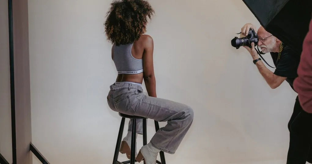 Photographer shooting a seated subject on a stool against a white studio backdrop with a softbox overhead, a typical portrait photography setup