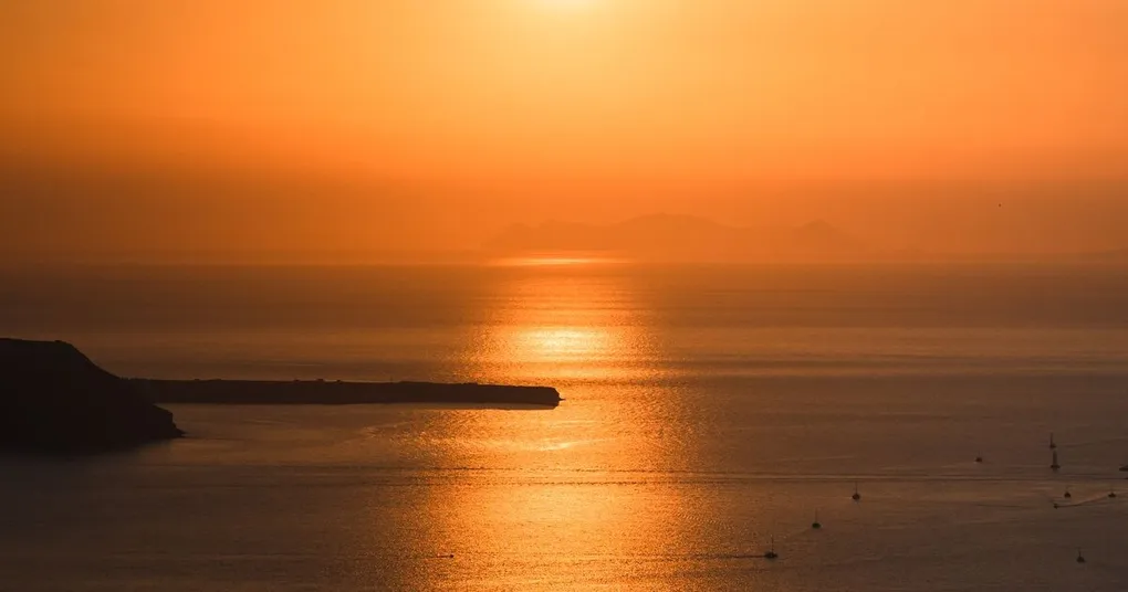 Sun low over the sea during golden hour, with an orange sky and a headland in silhouette