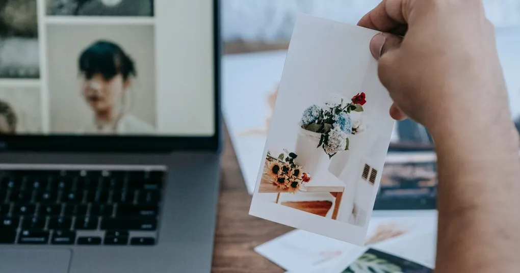 Hand holding a printed photo in front of a laptop displaying image files, comparing print and screen resolution