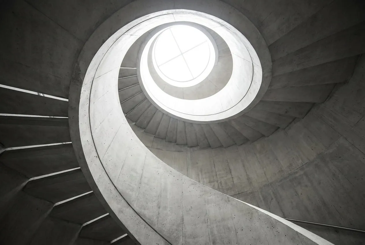 A spiral staircase photographed from below, the railing forming a diagonal leading line that sweeps toward a bright circular skylight