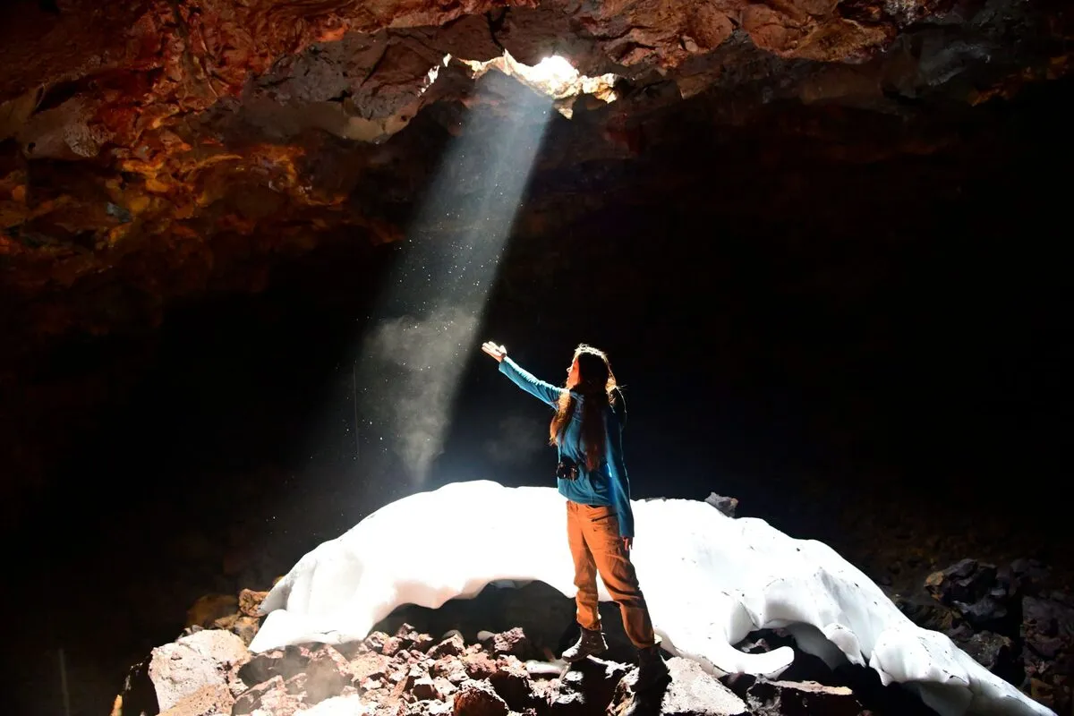 Person standing on snow inside a dark cave, reaching toward a bright shaft of daylight from an opening overhead