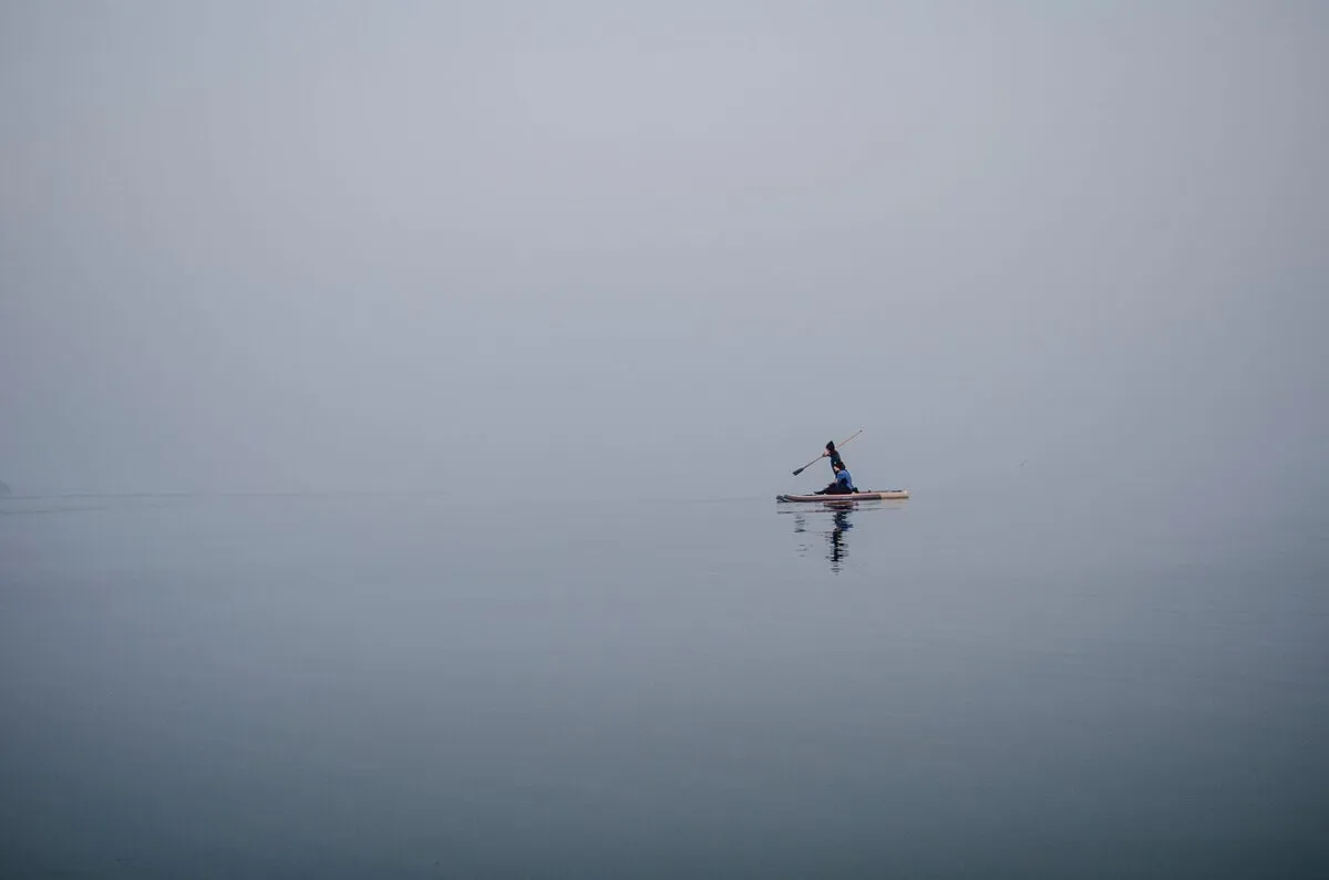 Lone paddler on flat gray water where mist erases the horizon into a single smooth field