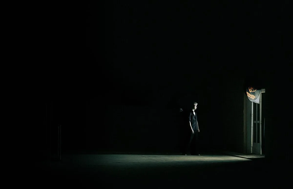 Figure standing in a small pool of light, surrounded by near-total darkness